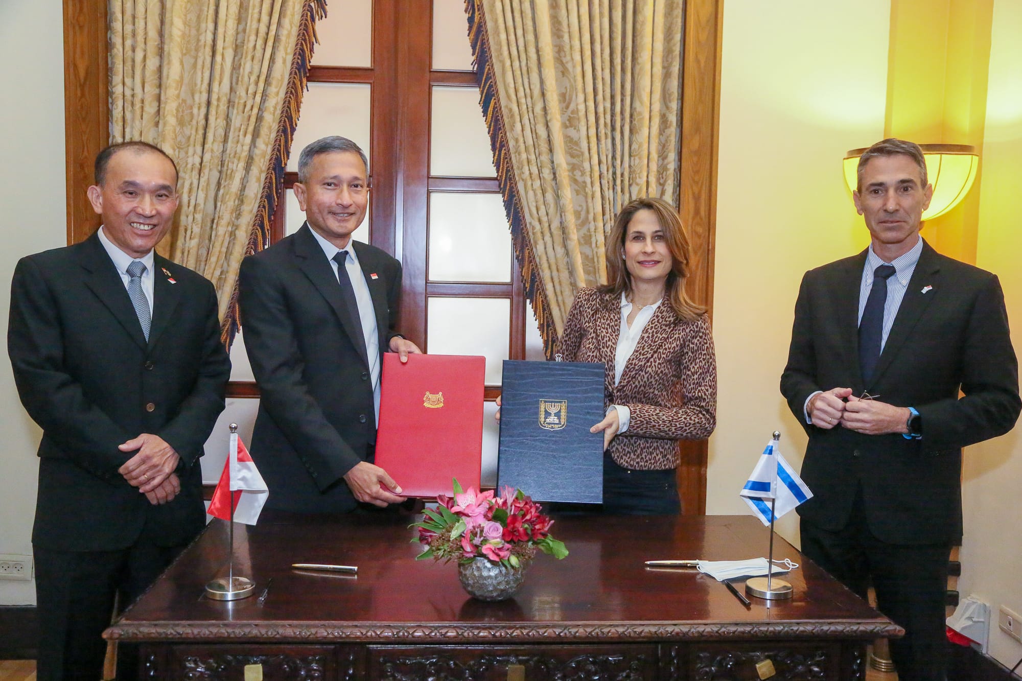 Four people in business attire holding red and blue folders, with flags on table.